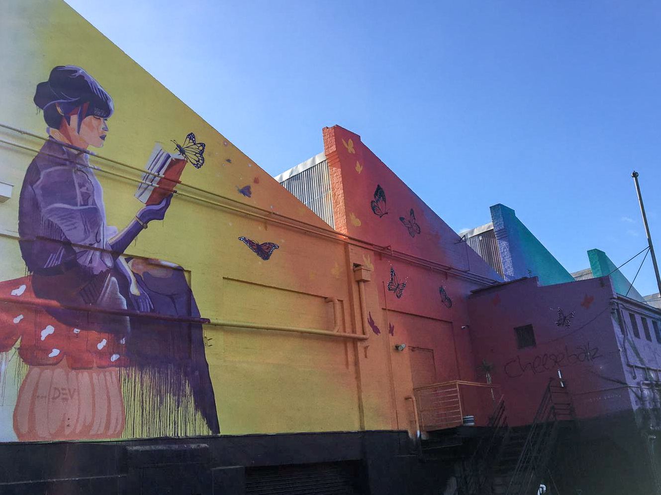 Exterior photo of building with street art of a women dressed in purple sitting on a red and white mushroom reading a book with a yellow butterfly. The wall turns gradually turns from yellow through the rainbow.