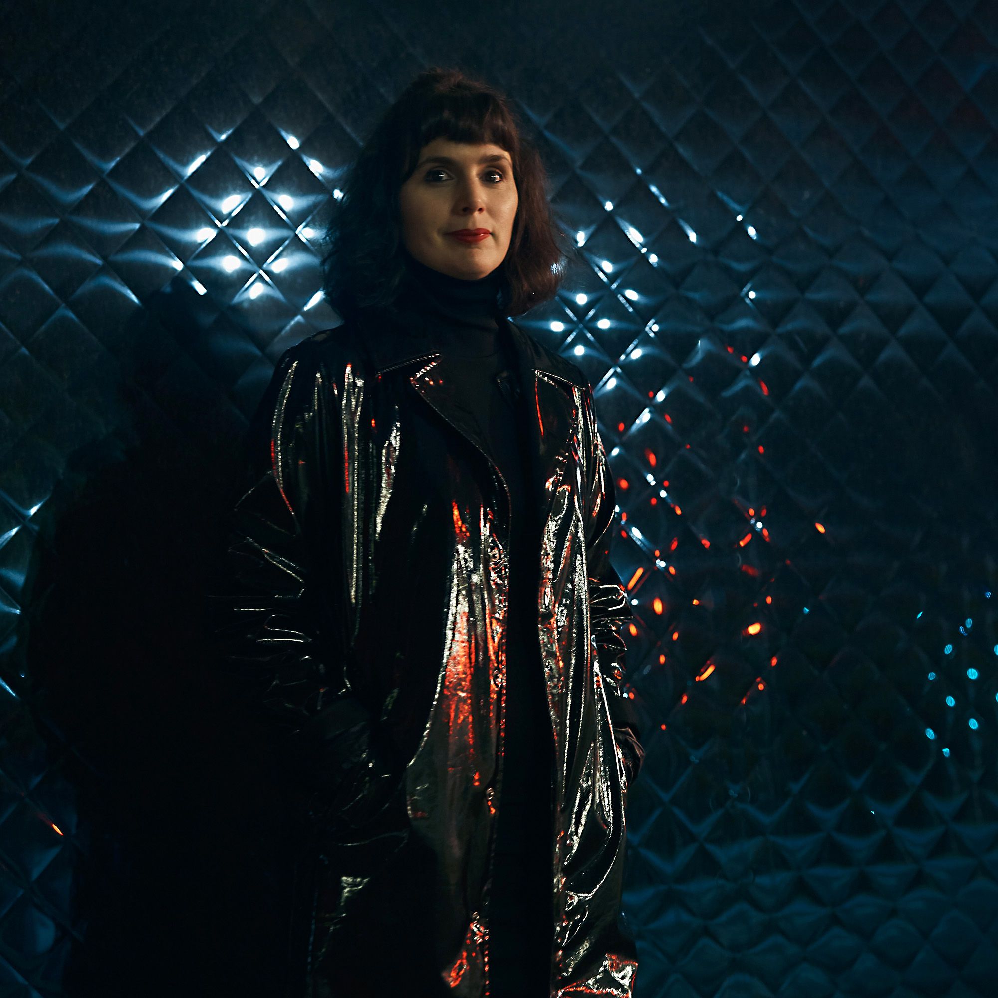 photo of a women with black hair standing in a shiny black nylon coat against a black leather wall