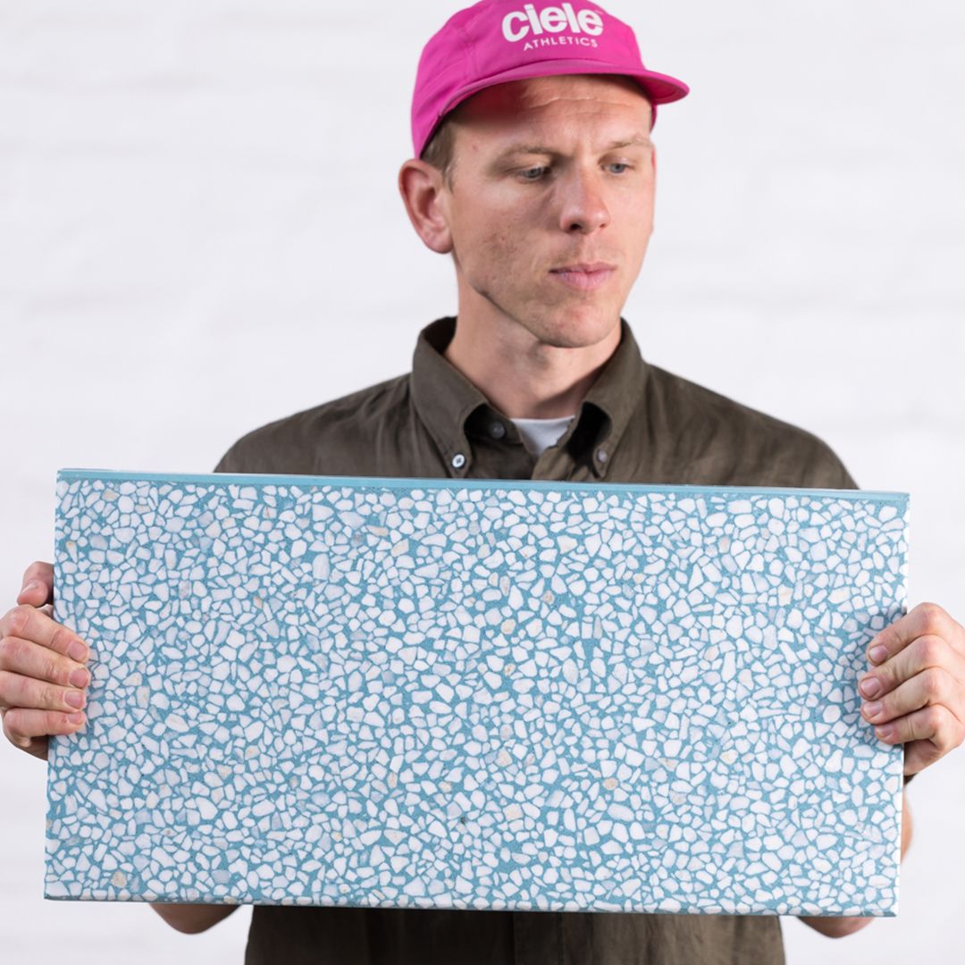 Photo of man holding blue terrazzo piece of concrete, wearing pink hat and green shirt.