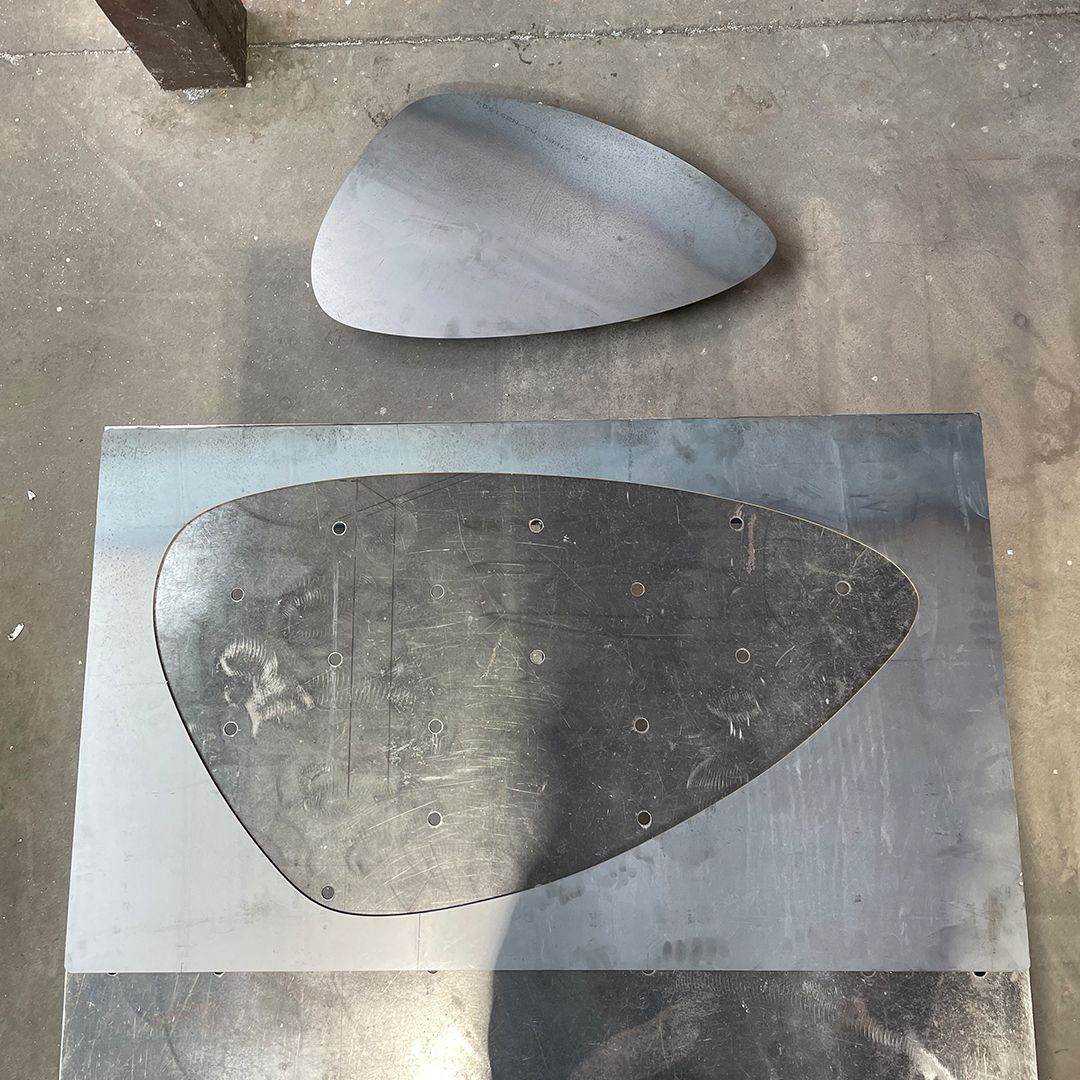 Offcut of aluminium sheet in a organic oval shape, the offcut lying next to sheet on concrete floor.